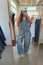 Load image into Gallery viewer, A woman takes a mirror selfie in a clothing store wearing light-wash blue denim overall shorts over a red and white striped t-shirt, paired with a white baseball cap.