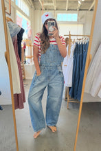 Load image into Gallery viewer, A woman takes a mirror selfie in a clothing store wearing light-wash blue denim overall with a red and white striped t-shirt and a white bucket hat with a red cherry.
