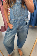 Load image into Gallery viewer, A person wearing light-wash blue denim overall jumpsuit or bib pants, photographed in a clothing store mirror. The garment has a faded wash, wide legs, and front pockets.