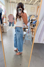Load image into Gallery viewer, A woman takes a mirror selfie in a boutique, wearing light-wash wide-leg cuffed jeans and a white tank top. The boutique interior has a concrete floor, clothing racks, and a wooden-framed mirror.