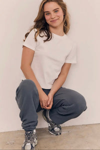 Person wearing a white t-shirt and gray sweatpants sitting against a plain wall.