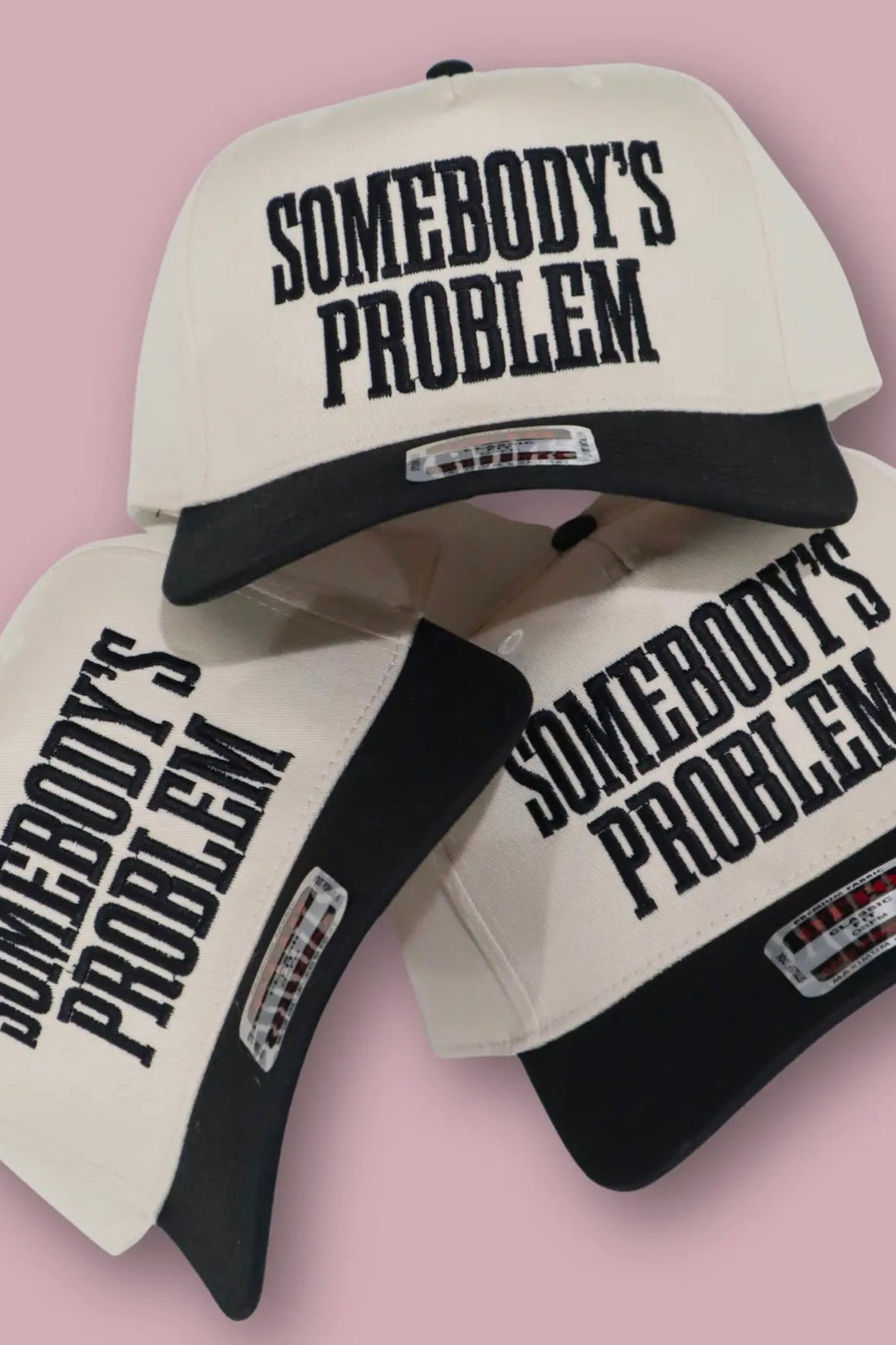 Three 'Somebody's Problem' caps on a pink background