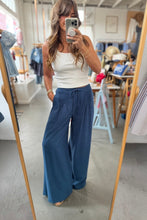 Load image into Gallery viewer, A woman takes a mirror selfie in a clothing store, wearing a white ribbed tank top and high-waisted, wide-leg blue pants with a drawstring waist.