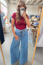 Load image into Gallery viewer, A woman takes a mirror selfie in a clothing boutique, wearing a short-sleeve red button-up top and high-waisted, wide-leg blue denim pants with a tie waist.
