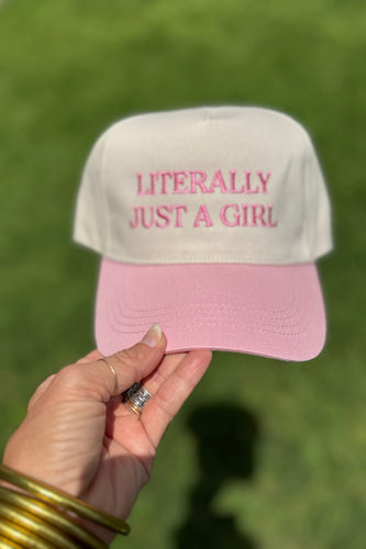 Pink and beige cap with 'Literally Just a Girl' text held by a hand against a blurred green background
