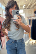 Load image into Gallery viewer, Woman taking a mirror selfie in a clothing store wearing a gray t-shirt and blue jeans.