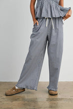 Load image into Gallery viewer, A person wearing loose wide-leg pants with a small black-and-white gingham pattern, a matching peplum top, and tan slip-on shoes, standing against a plain light gray wall.