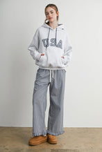 Load image into Gallery viewer, A woman stands in a studio wearing a light gray hoodie with 'USA' printed across the chest, paired with wide-leg gray-and-white gingham pants and tan platform mule shoes.