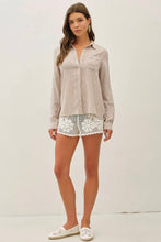Load image into Gallery viewer, A woman modeling a light beige linen button-up shirt with a chest pocket, paired with white floral lace shorts and white slip-on shoes, against a plain off-white studio background.