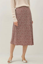 Load image into Gallery viewer, A model wearing a knee-length A-line skirt with a dense small-scale floral or ditsy print in dusty rose, burgundy, and cream, styled with a beige knit sweater and white heeled sandals against a light gray studio background.