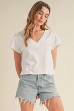 Load image into Gallery viewer, A woman wearing a white short-sleeve V-neck t-shirt and light-wash distressed denim shorts, standing against a plain beige studio background.