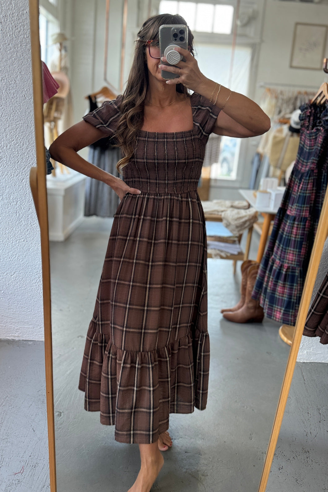 Woman taking a mirror selfie wearing a brown plaid dress in a store.