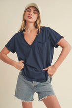 Load image into Gallery viewer, A woman modeling a navy blue short-sleeve V-neck t-shirt with a chest pocket, paired with light-wash denim shorts and a beige baseball cap, against a solid cream background.