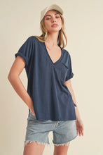Load image into Gallery viewer, A woman wearing a navy blue short-sleeve V-neck t-shirt with rolled cuffs and a chest pocket, paired with light-wash denim shorts and a beige baseball cap, against a solid cream background.