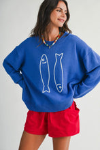 Load image into Gallery viewer, A woman wearing a bright blue crewneck sweater with white fish line drawings on the front, paired with red shorts, against a light gray studio background.
