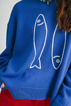 Load image into Gallery viewer, A close-up of a person wearing a bright blue knit sweater with large white fish line drawings on the front, paired with red bottoms against a light neutral background.