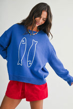 Load image into Gallery viewer, A woman wearing a bright blue knit sweater with two white fish line drawings on the front, paired with red shorts, photographed against a plain light gray studio background.