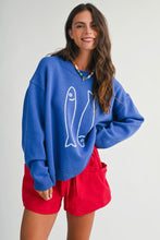 Load image into Gallery viewer, A woman wearing a vibrant blue knit sweater with a white fish graphic and bright red shorts, standing against a light gray studio background.