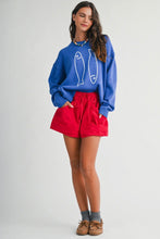 Load image into Gallery viewer, A woman modeling a blue crewneck sweatshirt with a white fish graphic, paired with bright red shorts, gray socks, and brown shoes, against a light gray studio background.