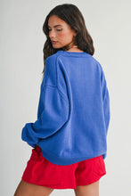 Load image into Gallery viewer, A woman wearing an oversized blue knit sweater and bright red shorts, photographed from the back against a plain light gray studio background.
