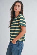 Load image into Gallery viewer, A woman wearing a green and yellow striped short-sleeve t-shirt with blue jeans, standing against a light gray studio background.