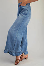 Load image into Gallery viewer, A person wearing light blue wide-leg denim jeans with a ruffled hem, paired with brown heeled sandals, shown from the waist down against a plain light gray studio background.