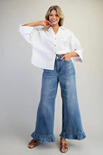 Load image into Gallery viewer, A woman modeling a white button-up shirt and high-waisted, wide-leg blue denim jeans with ruffled hems, paired with brown sandals, against a neutral light gray studio background.