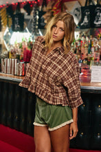Load image into Gallery viewer, A woman posing in a rustic bar setting, wearing a brown and beige plaid ruffled shirt and olive green velvet shorts with white lace trim.