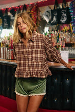Load image into Gallery viewer, A woman stands in a bar setting wearing a brown and beige plaid button-up shirt with a ruffled hem and green velvet shorts with a white lace trim.
