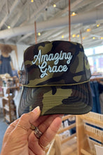 Load image into Gallery viewer, A camouflage trucker-style cap with 'Amazing Grace' embroidered in white script on the front, held by a hand inside a retail store.