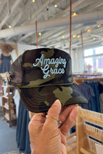 Load image into Gallery viewer, A hand holding a camouflage trucker hat with 'Amazing Grace' embroidered in white script on the front, set against a blurred clothing store background.