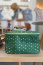 Load image into Gallery viewer, A green Kate Spade New York lunch bag with a white polka dot pattern, featuring a top zipper, carrying handle, and white script text, displayed on a light wooden table in a blurred boutique setting.