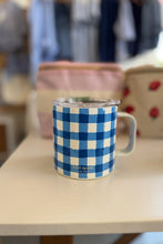 Load image into Gallery viewer, A Kate Spade enamel mug with a blue and white gingham pattern, sitting on a light-colored tabletop with a blurred background showing other bags.