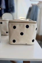Load image into Gallery viewer, A cream canvas cosmetic bag with black polka dots and a gold zipper, sitting on a white table in a softly blurred boutique setting.