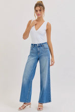 Load image into Gallery viewer, A woman modeling a pair of high-waisted, wide-leg blue denim jeans with a white sleeveless top and white heeled sandals, standing against a plain light gray studio background.