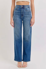 Load image into Gallery viewer, A model wearing a pair of high-waisted, wide-leg blue denim jeans with a faded wash, paired with a beige crop top and clear high-heel sandals, set against a plain light gray studio background.