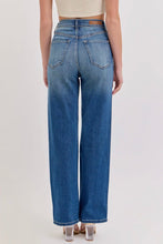Load image into Gallery viewer, A person shown from the back wearing high-waisted, wide-leg blue denim jeans with a tan leather patch on the back waistband, paired with a beige crop top and clear high heels against a light gray studio background.