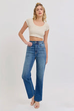 Load image into Gallery viewer, A woman modeling a pair of high-waisted, wide-leg blue jeans with a cream-colored crop top against a plain light gray studio background.