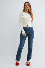 Load image into Gallery viewer, A woman modeling a cream ribbed knit cropped cardigan, blue straight-leg jeans with red zipper details at the cuffs, and red shoes, standing against a light gray studio background.