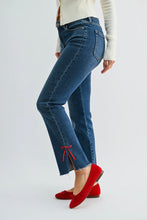 Load image into Gallery viewer, A person wearing blue cropped flare jeans with a raw hem and red side bow details, paired with a white ribbed sweater and red flats, shown from the waist down against a light gray studio background.