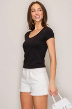 Load image into Gallery viewer, A woman modeling a black short-sleeve scoop neck t-shirt paired with white denim shorts, holding a white handbag against a plain light gray background.