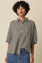 Load image into Gallery viewer, A woman wearing a black and white gingham checkered short-sleeve button-up shirt with a chest pocket, paired with blue denim jeans, against a solid beige background.