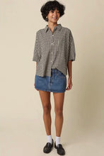 Load image into Gallery viewer, A woman modeling a black and white gingham short-sleeve button-up shirt, paired with a blue denim mini skirt, white socks, and black loafers, against a solid beige studio background.