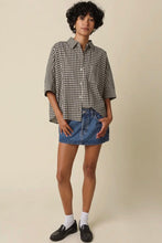 Load image into Gallery viewer, A model stands against a plain beige studio background wearing a black and white gingham short-sleeve button-up shirt, a blue denim mini skirt, white socks, and black loafers.