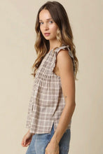 Load image into Gallery viewer, A woman wearing a sleeveless beige plaid top with ruffled shoulder details and blue jeans, standing against a solid beige background.
