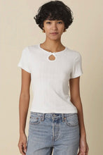 Load image into Gallery viewer, A woman modeling a white short-sleeve ribbed knit top with a circular cutout at the neckline, paired with light blue jeans, against a solid beige background.