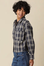 Load image into Gallery viewer, A woman with short curly black hair wearing a navy blue and white plaid button-up shirt and blue jeans, standing against a solid beige background.