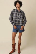 Load image into Gallery viewer, A woman modeling a long-sleeve navy and white plaid button-down shirt, paired with denim shorts and brown leather cowboy boots, against a solid beige studio background.