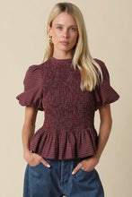 Load image into Gallery viewer, A woman modeling a smocked short-sleeve blouse with a red and black gingham pattern, paired with blue jeans, against a solid beige background.