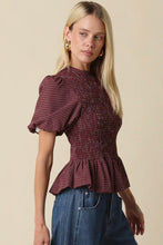 Load image into Gallery viewer, A blonde woman wearing a dark red and black checkered puff-sleeve peplum top with blue jeans, standing against a solid beige background.